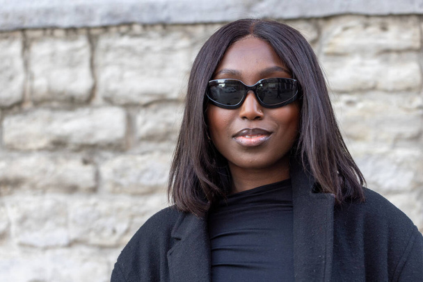 Deze foto legt een Afro-Amerikaanse vrouw vast die vertrouwen en stijl uitstraalt, met een chique zwarte jas over een coltrui, gecombineerd met een verfijnde zonnebril. Haar gecomponeerde uitdrukking tegen de - Foto, afbeelding