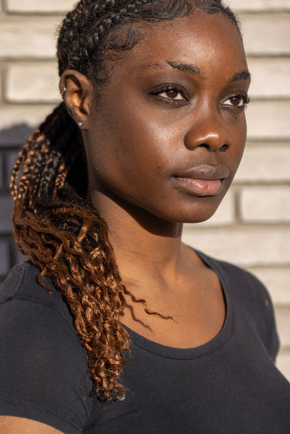 Αυτό το πορτραίτο αιχμαλωτίζει μια νεαρή Αφρικανή που ατενίζει την απόσταση, φωτισμένη από το ζεστό φως του ήλιου που λούζει τη μία πλευρά του προσώπου της. Τα πλεκτά μαλλιά της, στολισμένα με τόνους καραμέλας, καταρράκτες πάνω - Φωτογραφία, εικόνα