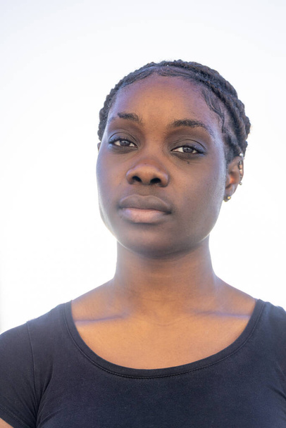 Dit is een frontaal portret van een Afrikaanse vrouw met een serene uitdrukking tegen een felwitte achtergrond. Haar haar is gestileerd in nette vlechten die haar gezicht symmetrisch omlijsten. De zachte verlichting wast - Foto, afbeelding