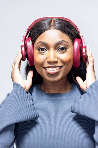This is a portrait of a cheerful young African woman with black hair, wearing a blue long-sleeved top and adjusting her red headphones. The headphones are clearly a significant feature, indicating a - Photo, Image