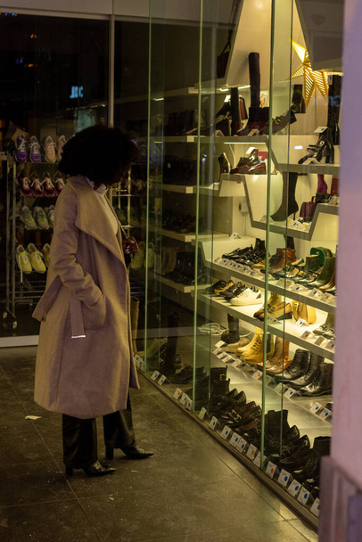 Bu görüntü vitrinlere bakan bir kadını görüntüler. Bir ayakkabı mağazasının önünde dururken, vitrine dalmış, silueti cama yansımış gibi görünüyor. Sıcaklık - Fotoğraf, Görsel