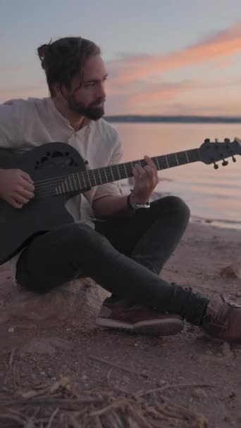 Spanischer Kaukasier spielt Gitarre am Strand am See bei Sonnenuntergang - Vertikales FullHD-Video - Filmmaterial, Video