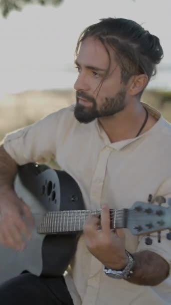 Красавчик-гитарист, играющий на классической акустической гитаре и поющий песню, глядя на объектив камеры в сельской местности Испании на закате - Vertical FullHD video - Кадры, видео