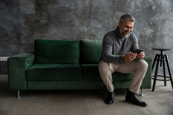 Un hombre está cómodamente sentado en un sofá verde, las piernas cruzadas mientras mira su teléfono celular, una pierna descansando sobre la rodilla de la otra - Foto, imagen