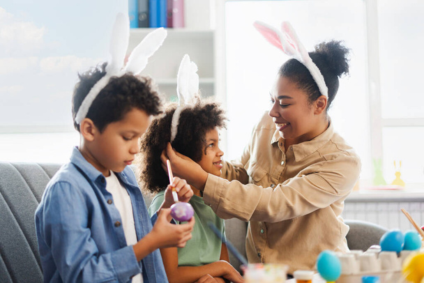 Madre sorridente e bambini carini con le orecchie da coniglio che dipingono e decorano le uova di Pasqua a casa. Felice celebrazione della famiglia afroamericana Pasqua insieme - Foto, immagini