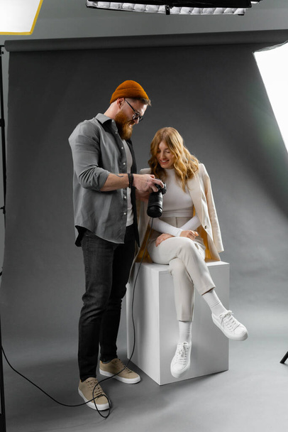 Un hombre está de pie junto a una mujer que está sentada en un cubo, mostrando el diseño de moda con ropa formal. La escena podría ser parte de un evento publicitario en una sala de exhibición de vehículos de lujo - Foto, imagen