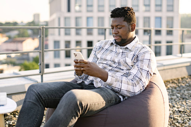 Cheerful African American person with smartphone resting in pouf chair on roof terrace during coffee break on mid day. Happy businesswoman in casual wear texting messages via mobile in open air. - Фото, зображення