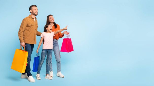 Shopping di famiglia. Sorridente papà caucasico, mamma e piccola figlia che trasportano borse shopper, indicando dito a parte lo spazio libero Mockup, raccomandare grande offerta su sfondo blu, Panorama - Foto, immagini