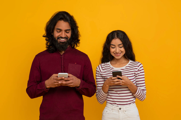 Fröhliches junges Paar aus dem Osten mit Smartphones in den Händen, die Seite an Seite auf gelbem Studiohintergrund stehen. Glückliche indische Männer und Frauen checken neueste mobile App - Foto, Bild