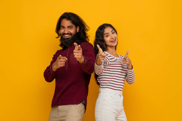 ポジティブな若いインド人男性と女性がカメラを指差します. ハッピーミレニアルヒンドゥーのカップルは,イエロースタジオの背景に隔離されたカジュアルな服のジェスチャーと笑顔を着ています - 写真・画像