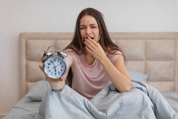 Drowsy young woman sits up in bed, stretching and yawning as she reluctantly presents an old-fashioned alarm clock, signaling the start of her day - Photo, Image