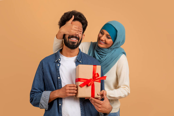 Verrassing voor jou. Romantische moslim vrouw bedekken echtgenoten ogen en geven cadeau, begroeten hem met verjaardag, trouwdag of Valentijnsdag, staan op beige studio achtergrond - Foto, afbeelding