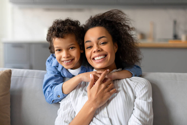 Feliz chico de pelo rizado negro con camisa azul abraza a su madre con alegría, ya que comparten un momento de amor juntos en un cómodo sofá y sonriendo a la cámara - Foto, imagen