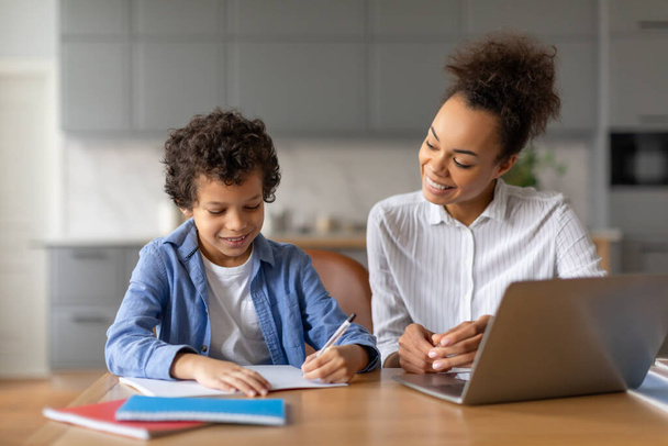 自然なカーリーヘアを持つ陽気な黒い母親は,彼が宿題を書くことに焦点を当て,自宅で教育環境を育成するために彼女の息子を導きます - 写真・画像