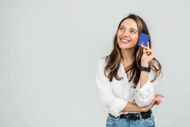 Jeune femme contemplative dans un chemisier blanc et un jean, tenant une carte bleue de crédit à son temple, avec une expression réfléchie et smartwatch, debout sur un fond gris - Photo, image