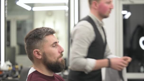 Un joven con una barba en una capa blanca se sienta en una silla en una barbería. El cliente está esperando un corte de pelo y afeitado de la barba 4k video. - Metraje, vídeo