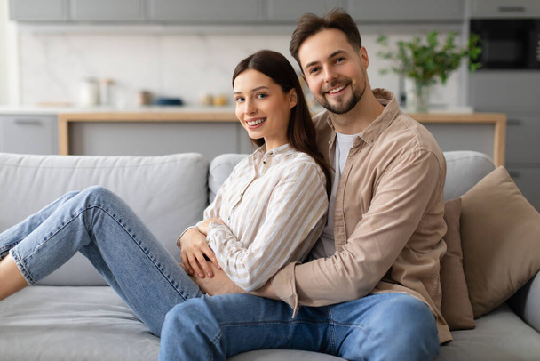 Usmívající se mladý pár sedí pohodlně na šedém gauči, objímají a užívají si spolu kvalitní čas v dobře osvětleném obývacím pokoji s moderním dekorem - Fotografie, Obrázek