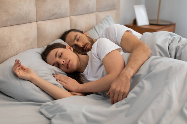 Peaceful couple in white t-shirts, embracing in restful slumber on cozy bed with soft pillows, symbolizing intimacy and comfort in serene bedroom - Photo, Image