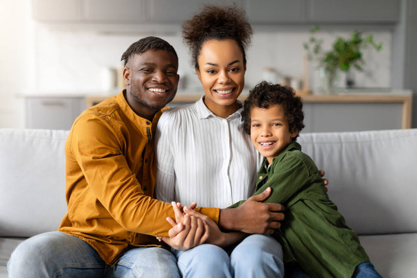 Cheerful african american family of three shares close, joyful moment, with bright smiles and embrace, exuding warmth and happiness in their home, sitting on sofa - Photo, Image