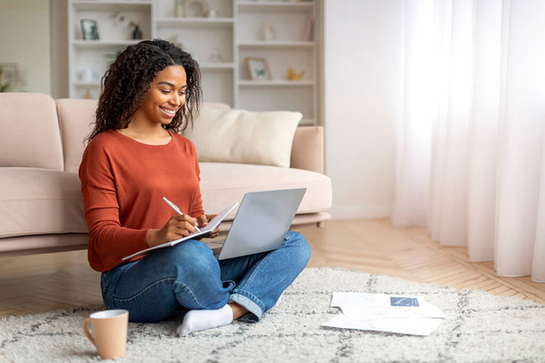 Νεαρή χαμογελώντας μαύρη γυναίκα χρησιμοποιώντας φορητό υπολογιστή στο σπίτι και λαμβάνοντας υπόψη, ευτυχισμένη αφρικανική αμερικανική γυναικεία μελέτη με υπολογιστή, ενώ κάθεται στο πάτωμα στο σαλόνι, απολαμβάνοντας σε απευθείας σύνδεση εκπαίδευση, αντιγραφή χώρου - Φωτογραφία, εικόνα