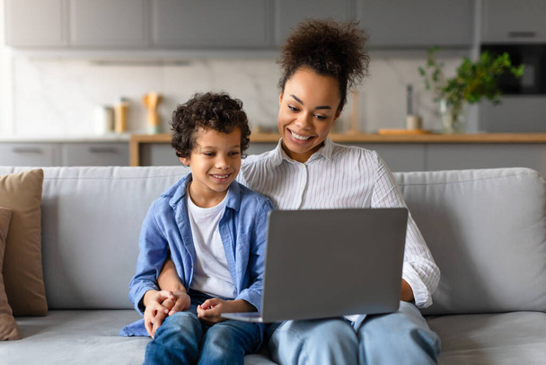 Mère noire souriante aux cheveux bouclés et fils en chemise bleue regarder ordinateur portable, profiter d'une activité partagée ensemble, choisir un dessin animé ou un film à regarder sur un canapé confortable salon - Photo, image
