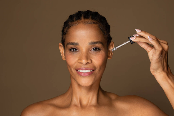 Mujer negra feliz de mediana edad usando gotero para aplicar suero facial, encarnando rutina de cuidado de la piel, sonriendo a la cámara, sobre fondo marrón suave - Foto, Imagen