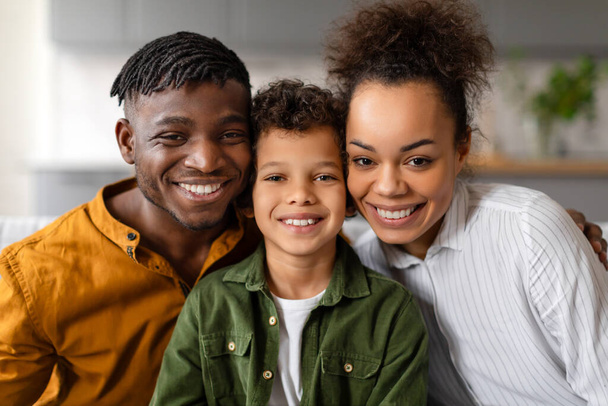 Vidám családi portré a fekete fiúról, akit szerető szülei ölelnek magukhoz, mindannyian szívből jövő mosollyal, meleg, szerető családdal, otthonos környezetben - Fotó, kép