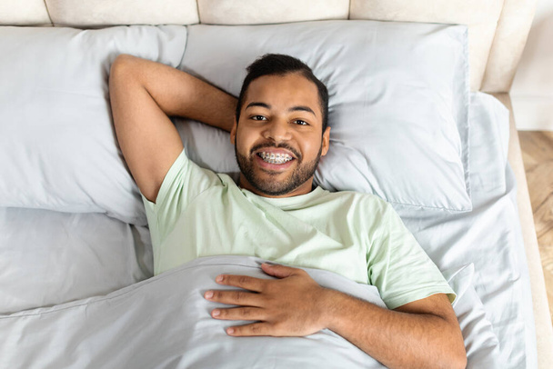 Positif beau jeune homme afro-américain barbu allongé au lit et souriant à la caméra, profitant d'une matinée ensoleillée, portant un pyjama, vue ci-dessus. Un sommeil et un mode de vie sains - Photo, image