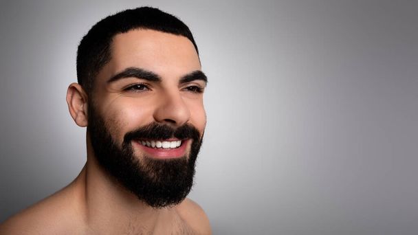 Foto de primer plano de hombre positivo guapo barbudo de Oriente Medio posando sobre fondo gris estudio, mirando espacio de copia para la publicidad y sonriente, panorama. Hombres cuidado de la piel concepto. - Foto, imagen