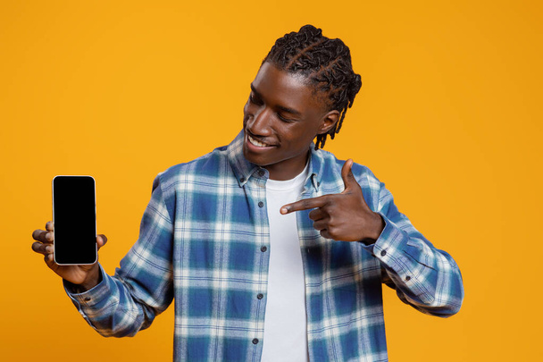Überprüfen Sie dies. Fröhlicher schwarzer Kerl, der auf leeres Smartphone in der Hand zeigt, lächelnder junger Afroamerikaner, der auf gelbem Studiohintergrund isoliert für eine neue mobile App wirbt, Mockup - Foto, Bild