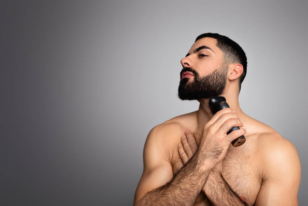 Beleza, higiene, barbear, arrumação e conceito de pessoas. Jovem do Oriente Médio olhando para o espaço de cópia e barba de barbear com aparador ou barbeador elétrico, isolado em fundo cinza - Foto, Imagem