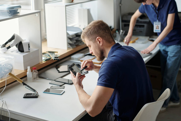 Młody pracownik biura napraw technicznych mocowanie drobnych szczegółów zdemontowanego telefonu komórkowego z małym śrubokrętem podczas siedzenia w miejscu pracy - Zdjęcie, obraz
