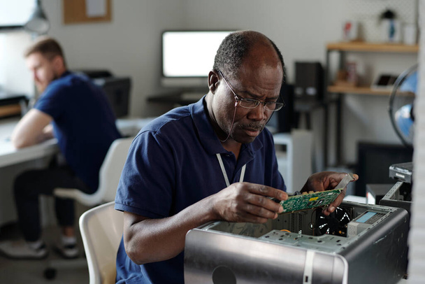 Riparatore afroamericano esperto in occhiali che guarda la scheda madre del computer e identifica i problemi tecnici in ufficio - Foto, immagini