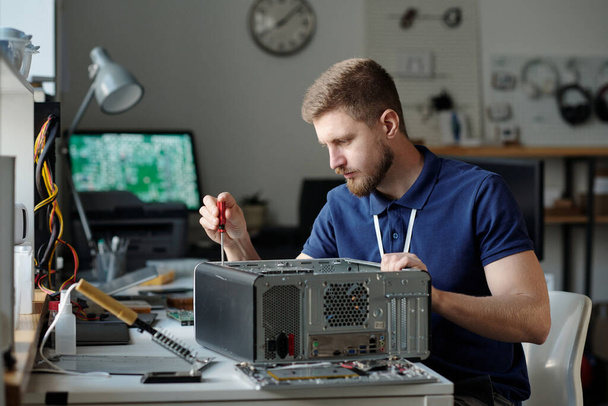 Jonge serieuze mannelijke technicus in donkerblauw uniform zittend op de werkplek in de reparatiedienst en los onderdelen van de computer processor - Foto, afbeelding