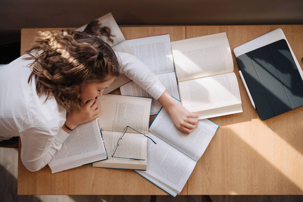 Una adolescente aparece abrumada, apoyando la cabeza en sus brazos en medio de un mar de libros de texto abiertos y una tableta - Foto, Imagen
