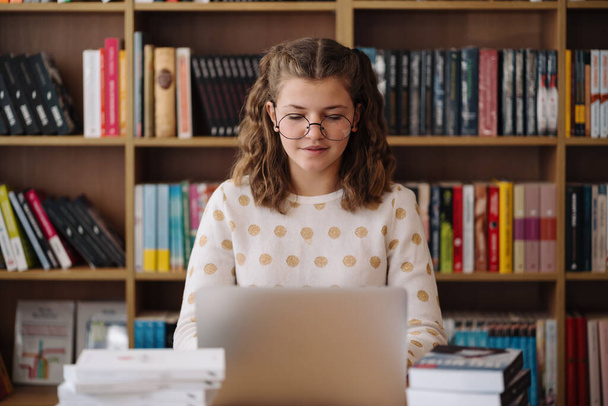 Tiener meisje met bril concentreert zich op haar laptop, studeren in een bibliotheek omringd door boeken - Foto, afbeelding