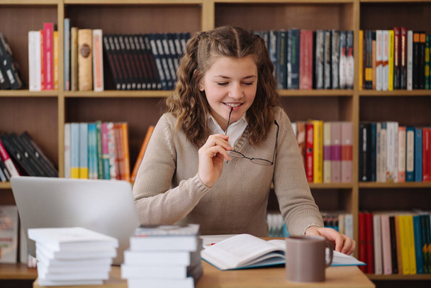 Egy figyelmes tinédzser lány szemüvegben elmélkedett a tananyagain a könyvtárban, körülvéve könyvekkel. - Fotó, kép