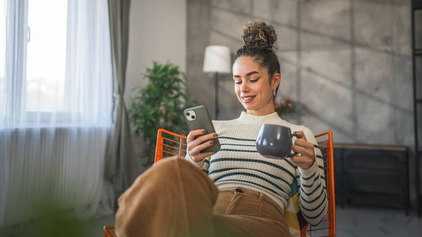 uma mulher adulto caucasiano feminino uso celular inteligente sms mensagens de texto enquanto sentar em casa segurar xícara de café - Foto, Imagem