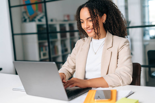 Positieve prachtige succesvolle zakenvrouw werken op laptop in kantoor, Braziliaanse zakenvrouw in formele jas zit aan een bureau in een moderne ruimte, werken aan een project in een laptop, glimlachen - Foto, afbeelding