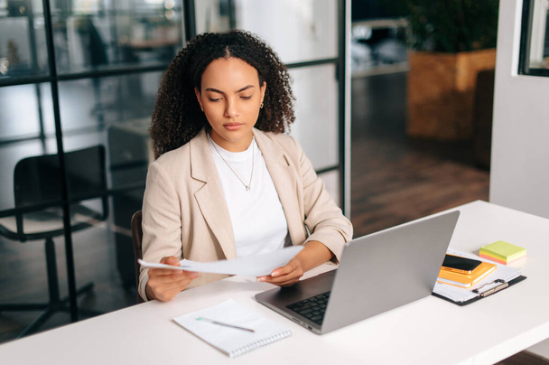 Serious confident brazilian or hispanic curly haired business woman, company ceo, financial manager, sitting at a work desk in modern office, working in a laptop, focused studying financial documents - Photo, Image
