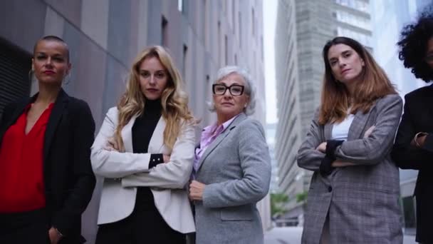 Низький кут руху жінок-підприємців у костюмах від розширеного жіночого бізнесу в місті. Група з п'яти серйозних колег різних рас і віків впевнено дивиться на камеру з обіймами, схрещеними на відкритому повітрі - Кадри, відео