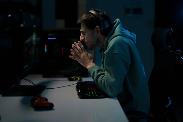 A pensive gamer with hands clasped, strategizing next moves in a dimly lit room - Photo, Image