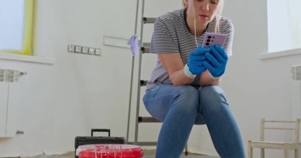 Merdivende oturan yorgun bir kadın, elinde boya silindiriyle telefonda konuşuyor, muhtemelen profesyonel yenileme yardımı istiyor. Yüksek kalite 4k görüntü - Video, Çekim