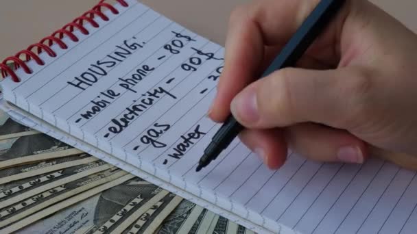 Mujer escribiendo en cuaderno gastos de vivienda. Dinero en efectivo en dólares Contando facturas de electricidad, gas, agua. Concepto de Consumo y Economía Eficientes. Hábitos de gasto - Metraje, vídeo