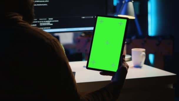 Programação desenvolvedor de software em home office, cruzando linhas de código entre tablet tela verde e tela do PC. TI admin segurando dispositivo portátil mockup durante a codificação no computador - Filmagem, Vídeo
