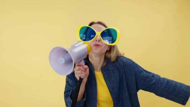 Aktive Frau in Comic-Brille hält Lautsprecher in der Hand und macht Werbung, während sie lacht. Energetische junge Frau unterhält die Menschen und erzählt lustige Witze im Megafon vor gelbem Hintergrund. - Filmmaterial, Video