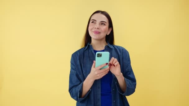 Dreamy caucasian woman looking aside with beaming smile on face while using modern smartphone in studio. Charming female millennial in denim shirt spending free time in social media with pleasure. - Footage, Video