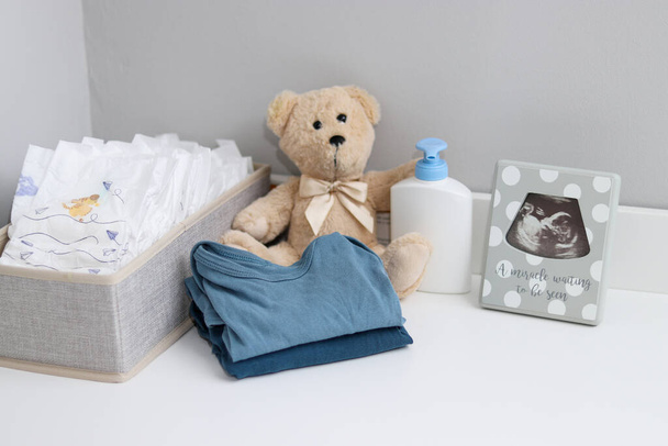 Een stapel luiers, rompertjes, teddybeer en babyspullen op witte commode, verpleegkamer  - Foto, afbeelding