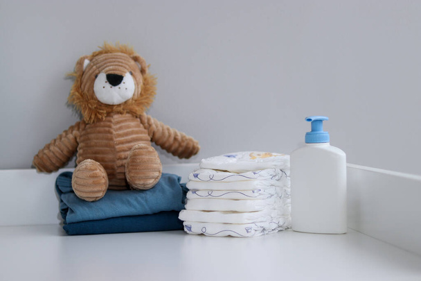 Een stapel luiers, blauwe rompertjes, pluche leeuwen en babyspullen op de commode, verpleegkamer - Foto, afbeelding