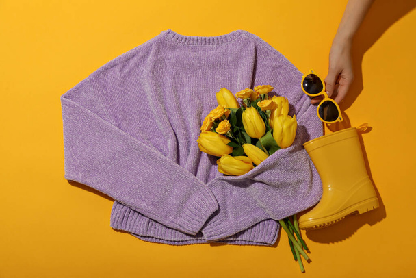Свитер, цветы, резиновые сапоги и очки в руке на желтом фоне, вид сверху - Фото, изображение